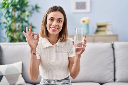 Téléchargez les photos : Hispanic woman drinking glass of water doing ok sign with fingers, smiling friendly gesturing excellent symbol - en image libre de droit