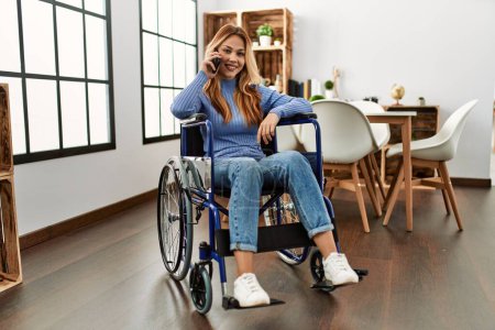 Téléchargez les photos : Jeune femme caucasienne parlant sur smartphone assis sur fauteuil roulant à la maison - en image libre de droit