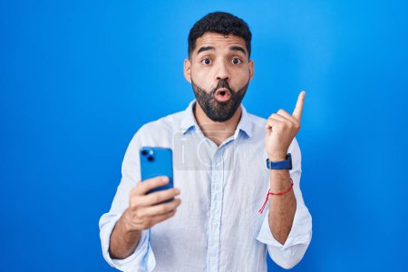 Téléchargez les photos : Homme hispanique avec barbe à l'aide d'un message de dactylographie smartphone surpris pointant du doigt sur le côté, bouche ouverte expression étonnée. - en image libre de droit