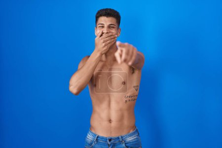 Foto de Young hispanic man standing shirtless over blue background laughing at you, pointing finger to the camera with hand over mouth, shame expression - Imagen libre de derechos