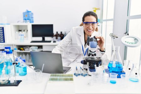 Téléchargez les photos : Jeune femme hispanique portant un uniforme de scientifique à l'aide d'un ordinateur portable et d'un microscope au laboratoire - en image libre de droit