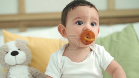 Photo for Adorable hispanic baby sucking pacifier sitting on bed at home - Royalty Free Image