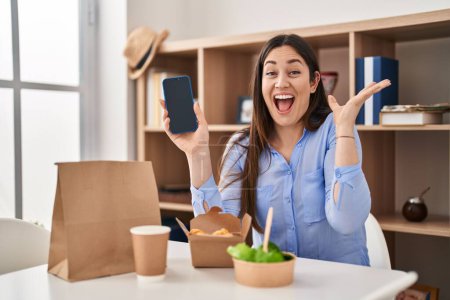 Téléchargez les photos : Jeune femme brune manger emporter de la nourriture à la maison montrant écran smartphone célébrant la victoire avec sourire heureux et expression gagnante avec les mains levées - en image libre de droit
