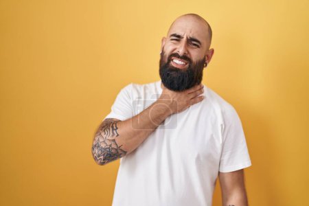 Foto de Young hispanic man with beard and tattoos standing over yellow background touching painful neck, sore throat for flu, clod and infection - Imagen libre de derechos