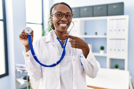 Téléchargez les photos : Médecin noir femme tenant stéthoscope souriant heureux pointant avec la main et le doigt - en image libre de droit