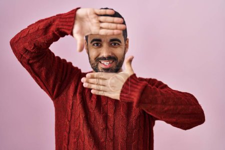 Téléchargez les photos : Jeune homme hispanique avec barbe portant pull décontracté sur fond rose souriant faisant cadre avec les mains et les doigts avec le visage heureux. concept de créativité et photographie. - en image libre de droit