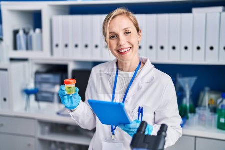 Téléchargez les photos : Jeune femme blonde scientifique utilisant un pavé tactile tenant un tube à essai d'urine au laboratoire - en image libre de droit