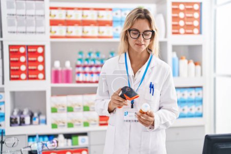 Téléchargez les photos : Jeune femme blonde pharmacien numérisation pilules bouteille à la pharmacie - en image libre de droit
