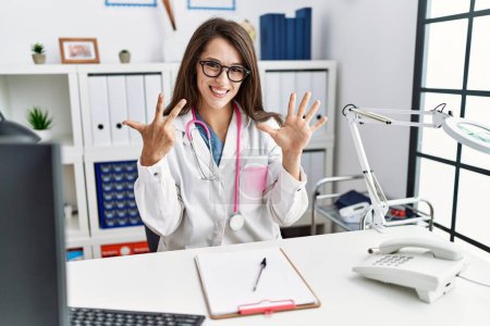 Foto de Joven doctora vistiendo uniforme médico y estetoscopio en la clínica mostrando y señalando con los dedos número ocho mientras sonríe confiada y feliz. - Imagen libre de derechos