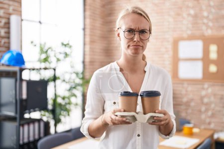 Foto de Mujer caucásica joven que trabaja en la oficina sosteniendo tazas de café escéptico y nervioso, frunciendo el ceño molesto debido a un problema. persona negativa. - Imagen libre de derechos
