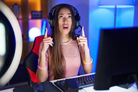 Photo for Young hispanic woman playing video games amazed and surprised looking up and pointing with fingers and raised arms. - Royalty Free Image
