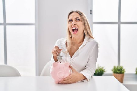 Photo for Young blonde woman holding piggy bank and house keys angry and mad screaming frustrated and furious, shouting with anger looking up. - Royalty Free Image