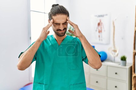 Photo for Young physiotherapist working at pain recovery clinic with hand on head for pain in head because stress. suffering migraine. - Royalty Free Image