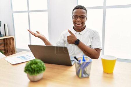 Téléchargez les photos : Jeune homme africain travaillant au bureau à l'aide d'un ordinateur portable montrant la main de la paume et faisant un geste correct avec les pouces levés, souriant heureux et joyeux - en image libre de droit