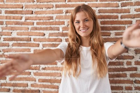 Photo for Young caucasian woman standing over bricks wall looking at the camera smiling with open arms for hug. cheerful expression embracing happiness. - Royalty Free Image