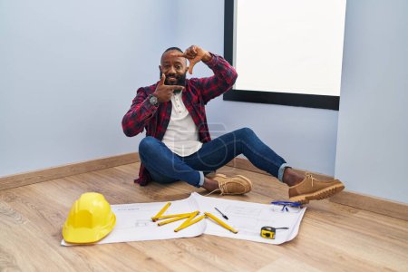 Photo for African american man sitting on the floor at new home looking at blueprints smiling making frame with hands and fingers with happy face. creativity and photography concept. - Royalty Free Image
