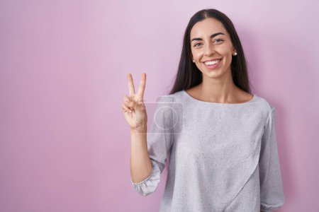 Téléchargez les photos : Jeune femme brune debout sur fond rose montrant et pointant vers le haut avec les doigts numéro deux tout en souriant confiant et heureux. - en image libre de droit