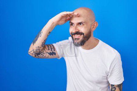 Photo for Hispanic man with tattoos standing over blue background very happy and smiling looking far away with hand over head. searching concept. - Royalty Free Image