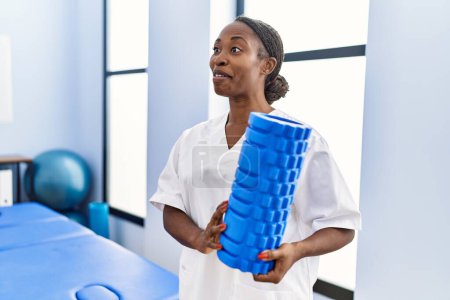 Foto de Mujer afroamericana fisioterapeuta sonriendo confiada sosteniendo rodillo de espuma en clínica de rehabilitación - Imagen libre de derechos