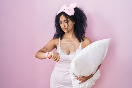Téléchargez les photos : Femme hispanique avec des cheveux bouclés portant un masque de sommeil et un pyjama tenant un oreiller vérifiant l'heure sur la montre-bracelet, détendue et confiante - en image libre de droit