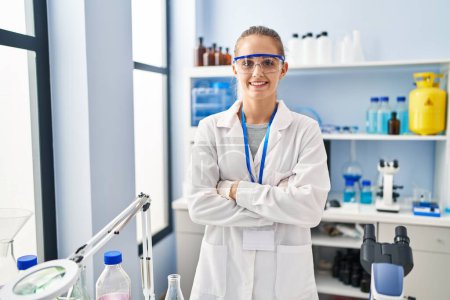 Foto de Mujer rubia joven vistiendo uniforme científico de pie con los brazos cruzados gesto en el laboratorio - Imagen libre de derechos
