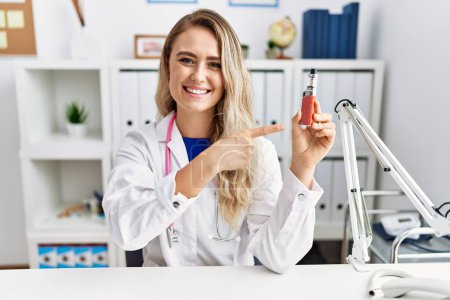 Téléchargez les photos : Jeune belle médecin femme tenant cigarette électronique à la clinique souriant heureux pointant avec la main et le doigt - en image libre de droit