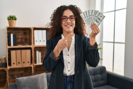 Photo for Young hispanic woman holding dollars smiling happy pointing with hand and finger - Royalty Free Image
