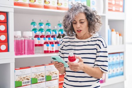 Téléchargez les photos : Middle age grey-haired woman client using smartphone holding medication bottle at pharmacy - en image libre de droit