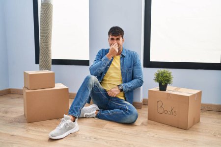 Téléchargez les photos : Jeune homme hispanique assis sur le sol à la nouvelle maison sentant quelque chose puant et dégoûtant, odeur intolérable, retenant souffle avec les doigts sur le nez. mauvaise odeur - en image libre de droit