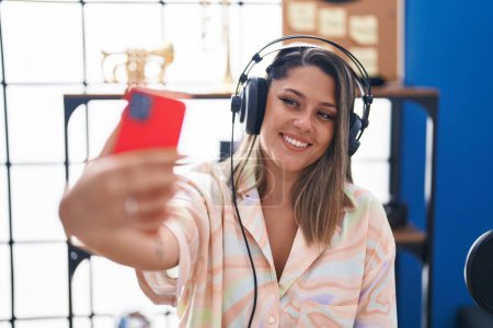 Photo for Young hispanic woman musician listening to music make selfie by smartphone at music studio - Royalty Free Image