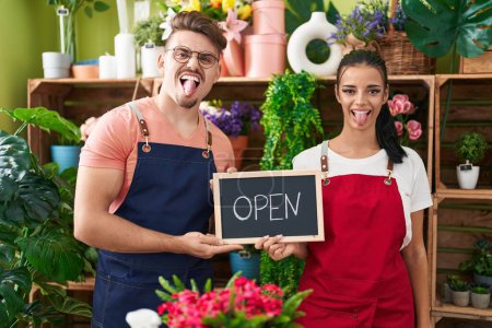 Photo for Young hispanic man a woman working at florist holding open sign sticking tongue out happy with funny expression. - Royalty Free Image