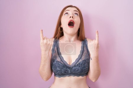 Foto de Redhead woman wearing lingerie over pink background amazed and surprised looking up and pointing with fingers and raised arms. - Imagen libre de derechos