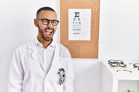 Téléchargez les photos : African american optician man standing by eyesight test angry and mad screaming frustrated and furious, shouting with anger. rage and aggressive concept. - en image libre de droit