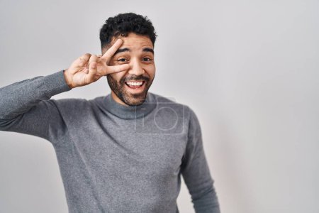 Photo for Hispanic man with beard standing over white background doing peace symbol with fingers over face, smiling cheerful showing victory - Royalty Free Image