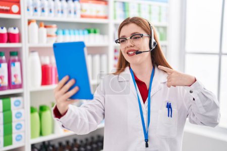 Téléchargez les photos : Redhead woman working at pharmacy drugstore doing video call with tablet smiling happy pointing with hand and finger - en image libre de droit