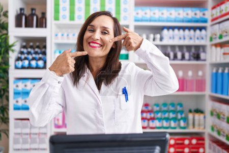 Téléchargez les photos : Middle age brunette woman working at pharmacy drugstore smiling cheerful showing and pointing with fingers teeth and mouth. dental health concept. - en image libre de droit