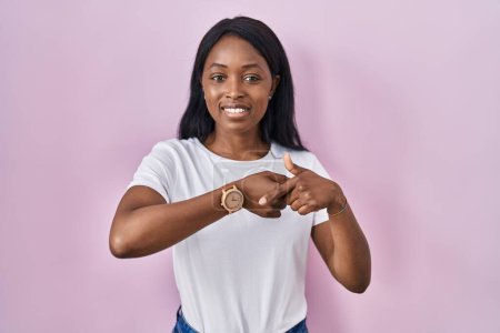 Foto de Mujer joven africana vistiendo camiseta blanca casual con prisa apuntando a ver el tiempo, la impaciencia, mirando a la cámara con expresión relajada - Imagen libre de derechos