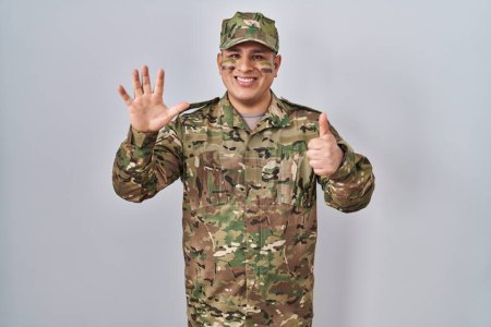 Téléchargez les photos : Jeune homme hispanique portant uniforme de camouflage de l'armée montrant et pointant vers le haut avec les doigts numéro six tout en souriant confiant et heureux. - en image libre de droit