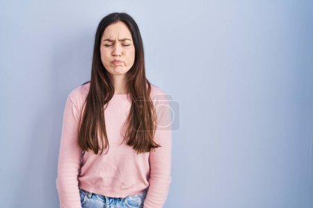 Photo for Young brunette woman standing over blue background puffing cheeks with funny face. mouth inflated with air, crazy expression. - Royalty Free Image