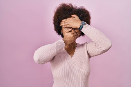 Téléchargez les photos : Jeune femme afro-américaine debout sur fond rose couvrant les yeux et la bouche avec les mains, surprise et choquée. cacher l'émotion - en image libre de droit