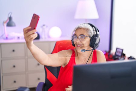 Foto de Streamer mujer de pelo gris de mediana edad sonriendo confiado hacer selfie por teléfono inteligente en la sala de juegos - Imagen libre de derechos