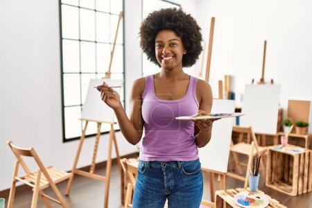 Foto de Joven mujer afroamericana sonriendo confiada sosteniendo el pincel y la paleta en el estudio de arte - Imagen libre de derechos