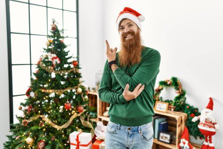 Téléchargez les photos : Homme rousse à longue barbe portant un chapeau de Noël par arbre de Noël avec un grand sourire sur le visage, pointant du doigt vers le côté en regardant la caméra. - en image libre de droit