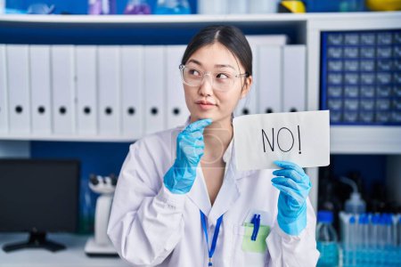 Photo for Chinese young woman working at scientist laboratory holding no banner serious face thinking about question with hand on chin, thoughtful about confusing idea - Royalty Free Image