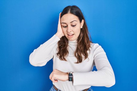 Téléchargez les photos : Jeune femme hispanique debout sur fond bleu regardant le temps de la montre inquiet, peur de se faire tard - en image libre de droit