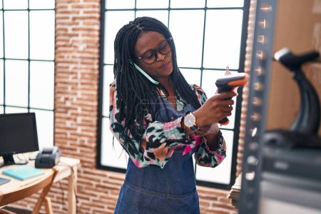 Foto de Mujer afroamericana comercio electrónico trabajador de negocios paquete de escaneo hablando en el teléfono inteligente en la oficina - Imagen libre de derechos