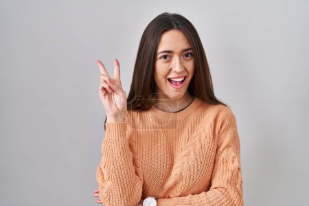 Photo for Young brunette woman standing over white background smiling with happy face winking at the camera doing victory sign with fingers. number two. - Royalty Free Image