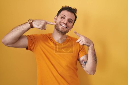 Téléchargez les photos : Jeune homme hispanique debout sur fond jaune souriant gai montrant et pointant avec les doigts dents et la bouche. concept de santé dentaire. - en image libre de droit