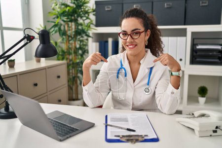 Photo for Young hispanic woman wearing doctor uniform and stethoscope looking confident with smile on face, pointing oneself with fingers proud and happy. - Royalty Free Image