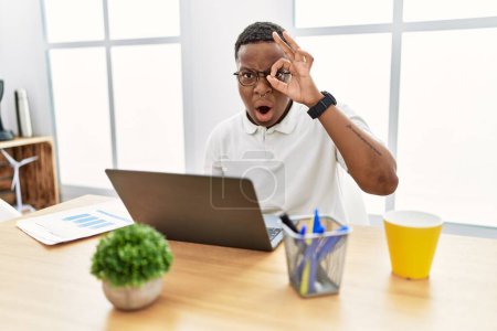 Foto de Un joven africano que trabajaba en la oficina usando un ordenador portátil haciendo un buen gesto sorprendió con la cara sorprendida, los ojos mirando a través de los dedos. expresión incrédula. - Imagen libre de derechos